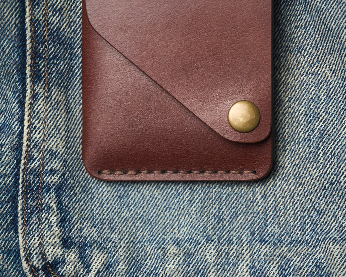 Close-up of a handcrafted leather wallet on denim, highlighting the thick hand-stitched thread, brass snap hardware, and rich brown leather grain