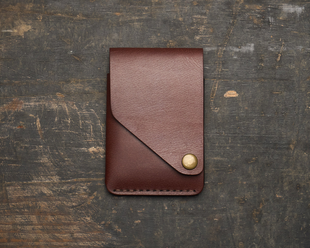 Front view of a minimalist brown, vegetable-tanned leather wallet with a distinctive angled flap and brass snap closure, resting on a rustic wooden surface.