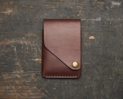 Front view of a minimalist brown, vegetable-tanned leather wallet with a distinctive angled flap and brass snap closure, resting on a rustic wooden surface.