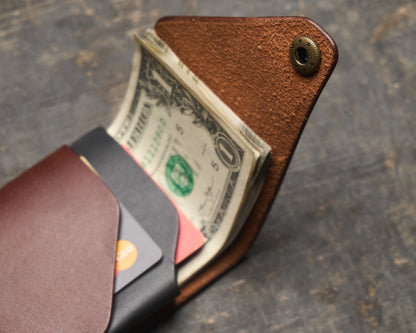 Close-up of an open, handmade leather wallet showing a brass snap, folded US dollar bills, and a contrasting black interior pocket holding a credit card