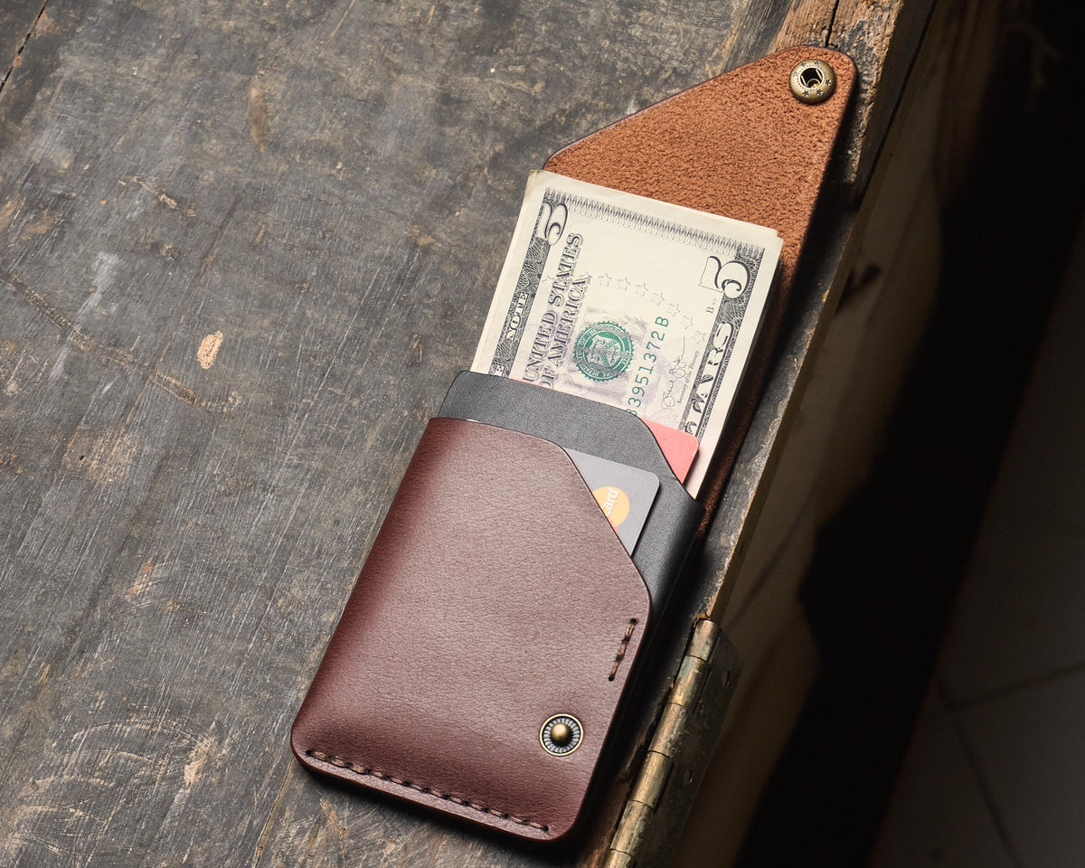 Open slim leather cash wrap wallet on a wooden desk, displaying interior black card slots filled with cards and folded five-dollar bills.