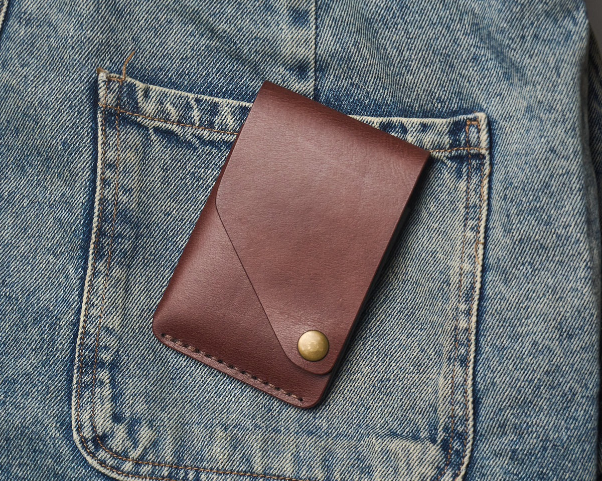 Minimalist brown leather front-pocket wallet with a secure snap closure resting inside the pocket of blue denim jeans.